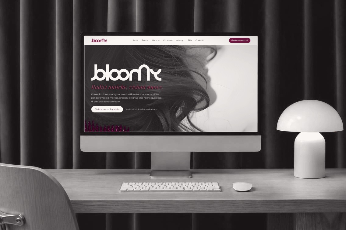 Monitor su una scrivania in legno con il sito di Studio Bloomy aperto, una lampada a fungo e una sedia, in bianco e nero con i dettagli bordeaux